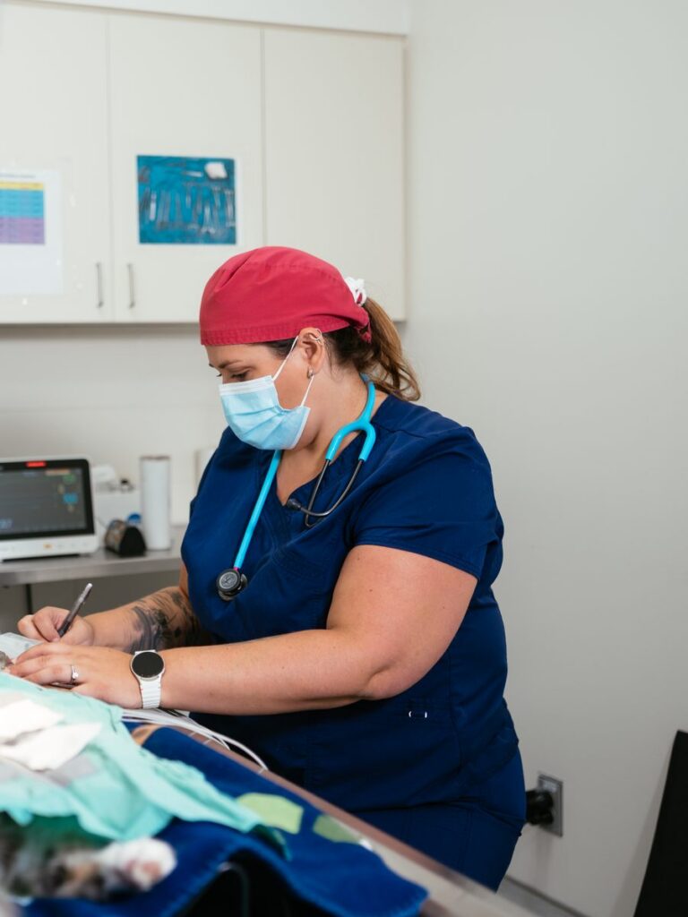 A woman in navy scrubs, a mask and a red surgical cap takes notes beside a cat under anesthesia.