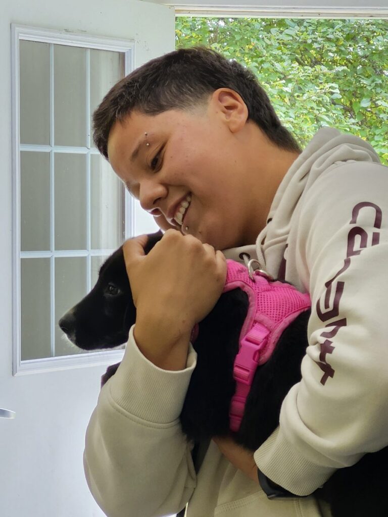 A smiling person with short dark hair hugs a black puppy in a pink harness close to their cheek.